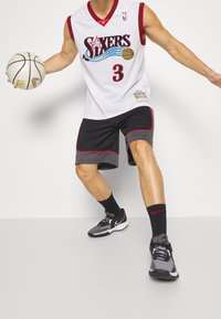 Mies, joka pitää Philadelphia 76ers -koripallopaidan ja shortseja, driblaa valkoista koripalloa, jalassaan Nike-tennarit ja mustat sukat, joissa on punainen logo.