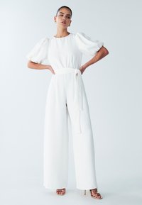 Witte jumpsuit met pofmouwen, een getailleerde taille en een wijde pijpvorm, gemaakt van gladde stof. Gecombineerd met sandalen met bandjes.