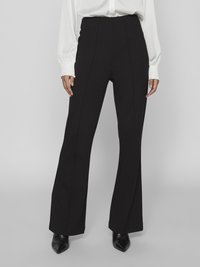 Black high-waisted trousers with flared legs, tailored design, and vertical seams. Worn with a white buttoned blouse and pointed black shoes.