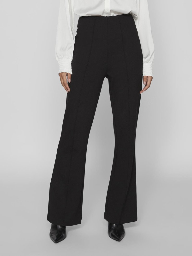 Black high-waisted trousers with flared legs, tailored design, and vertical seams. Worn with a white buttoned blouse and pointed black shoes.