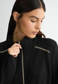 Črna zip-up jakna z visokim ovratnikom, okrašena z zlatimi zadrgami in elegantnim dizajnom. Gladka tekstura tkanine z minimalističnimi detajli.