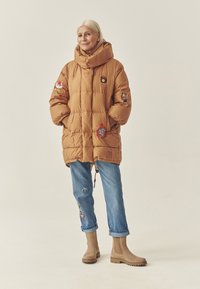 Brūns pūkains mētelis ar augstu apkakli un vairākiem plāksteriem, apvienots ar sarullētām zilo džinsiem un brūniem potīšu zābakiem. Strukturēta, silta auduma.