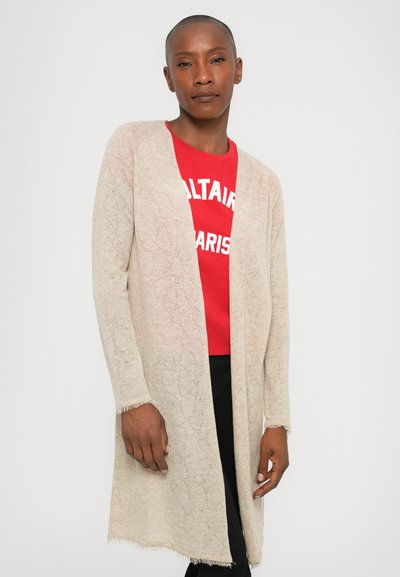 Light beige knitted cardigan with an open front, featuring a leaf pattern and fringe trim. Worn over a red graphic t-shirt and black pants.