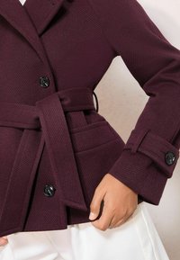 Bordo spalvos paltas su siaurėjančiu tekstūros raštu, su diržu juosmenyje, didelėmis juodomis sagomis ir šoniniais kišenėmis; turi įpjautą apykaklę ir pritaikytą kirpimą.