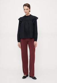 Blusa nera a pois con spalle arricciate, maniche lunghe e scollo alto. Abbinata a pantaloni dritti bordeaux e ballerine nere.