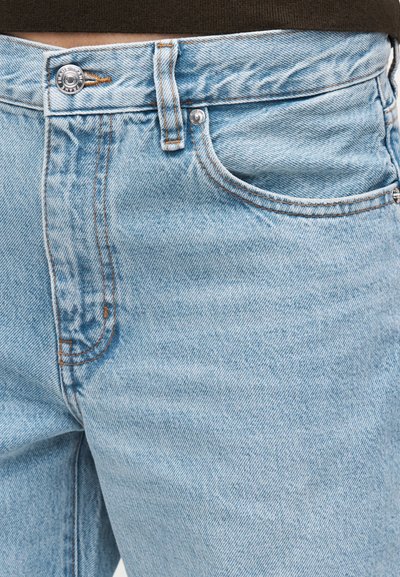 Φωτεινό γαλάζιο τζιν με μπροστινή τσέπη, θηλιά για ζώνη, μεταλλικό κουμπί και εμφανή ραφές που φοριέται από ένα άτομο με ορατό μέρος της μέσης.