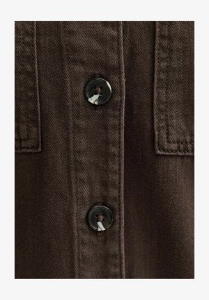 Nahaufnahme eines braunen Stoffes mit zwei gemusterten dunklen Knöpfen, die mit passendem Faden angenäht sind, zeigt detaillierte Nähte und strukturiertes Material.