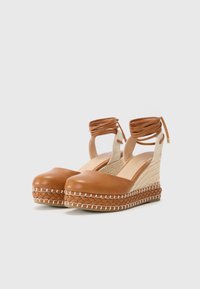 Alpargatas de cuña de cuero marrón con suela de yute trenzado y cintas que se envuelven alrededor del tobillo. Presentan una textura suave y un diseño de punta redondeada cerrada, con acentos en beige en contraste.