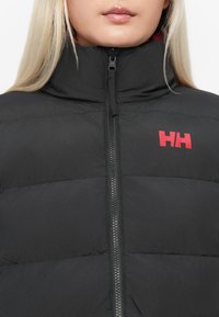 Chaqueta de plumas negra con cuello alto, cierre de cremallera frontal y logo rojo. Presenta un diseño acolchado y un estilo elegante y aerodinámico.