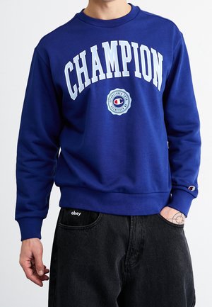 Person trägt einen blauen Champion-Sweatshirt mit weißer Schrift und Logo, kombiniert mit schwarzen Jeans, die ein kleines Tattoo an der rechten Hand zeigen.