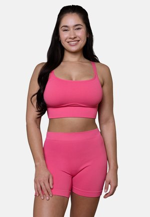 Junge Frau lächelnd, trägt ein passendes, geripptes pinkfarbenes Sport-BH und hochtaillierte Biker-Shorts vor schlichtem Hintergrund.