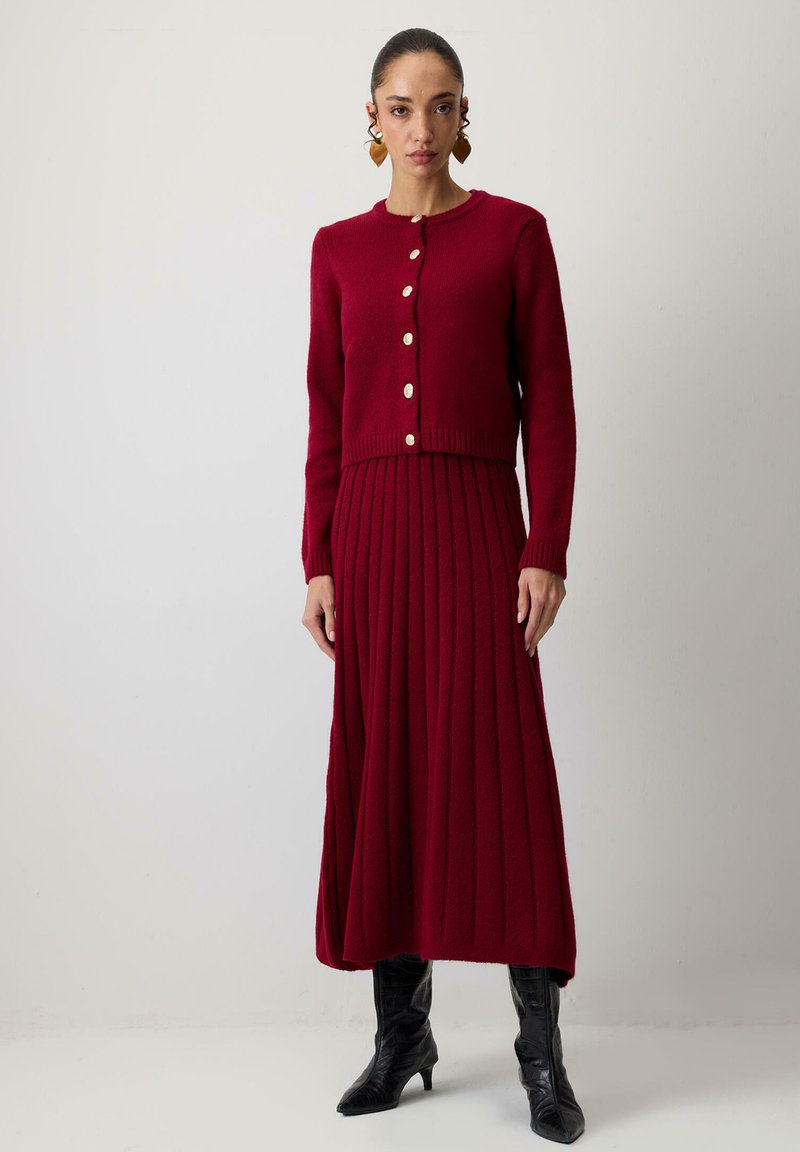 Zestaw sukienki z prążkowanego dzianiny i kardiganu w głębokim czerwonym kolorze, z okrągłym dekoltem, zapinany na guziki oraz fakturowaną tkaniną. Czarne botki na obcasie dopełniają look.