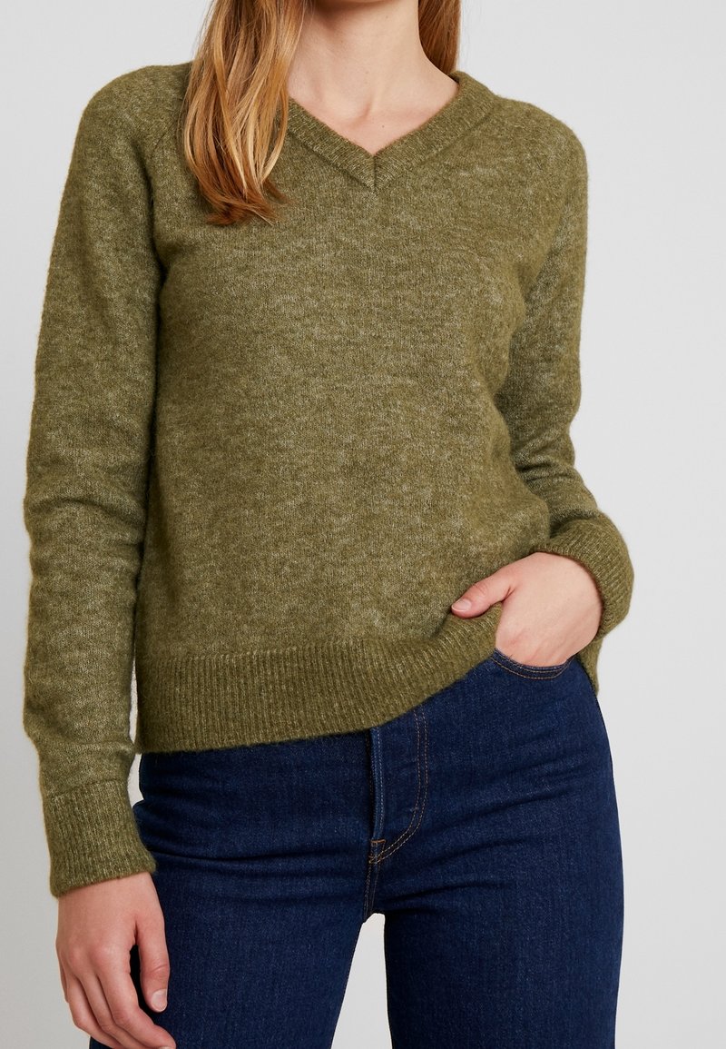 Jersey de punto verde oliva con escote en V, puños y dobladillo acanalados, hecho de una suave tela texturizada. Combinado con pantalones vaqueros azul oscuro.