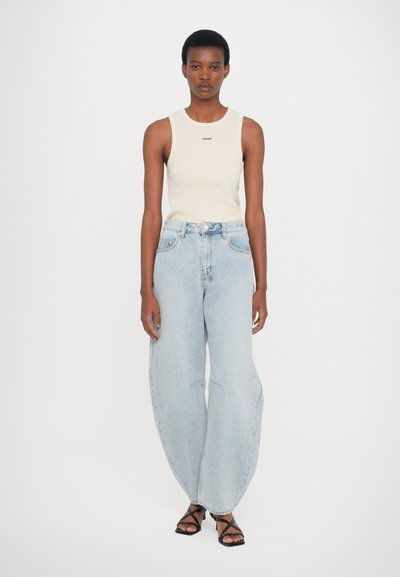 Lichtbeige aansluitend tanktop met een klein logo, gecombineerd met oversized lichtblauwe jeans met een ontspannen pasvorm en subtiele vervaging. Zwarte sandalen met bandjes.