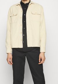 Giacca beige chiaro con chiusura a bottoni, due tasche sul petto e maniche lunghe, indossata sopra una camicia nera e abbinata a pantaloni scuri.