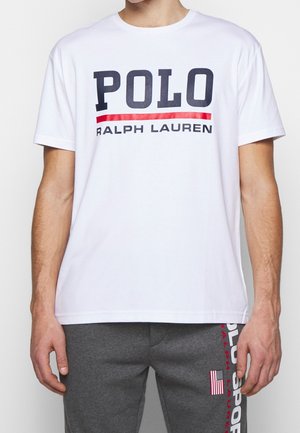 Mężczyzna w białej koszulce z napisem "POLO RALPH LAUREN" oraz szarych spodniach z logo Polo Sport i małą amerykańską flagą.