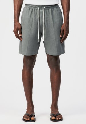 Man wearing gray textured shorts with white drawstrings and black flip-flops, standing against a plain white background.