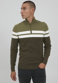 Chaki žalios spalvos zip-up megztinis su baltomis ir tamsiai žaliomis horizontaliomis juostomis, turintis tekstūruotą mezginį ir guma apdirbtas rankoves bei apačią.