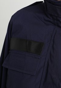 Veste bleu marine avec une sangle horizontale noire traversant une poche à rabat sur la poitrine, dotée d'un col montant.