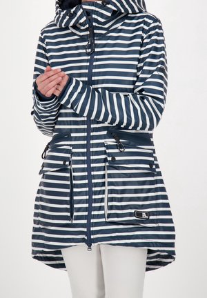 Imperméable à capuche rayé horizontal bleu marine et blanc avec fermeture éclair à l'avant, poches à rabat et cordons sur les manches et la capuche, porté avec un pantalon blanc.