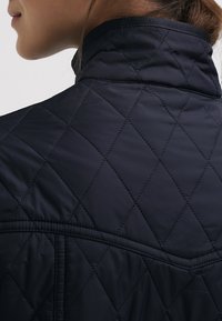 Personne portant une veste matelassée foncée avec des coutures en forme de losange, vue de dos et de côté, mettant l'accent sur le col et la zone des épaules.