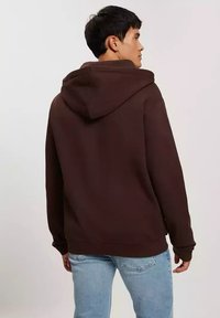 Brązowa bluza z kapturem wykonaną z teksturowanej tkaniny, związywana sznurkiem z kapturem i kieszenią kangurką, w połączeniu z jasnoniebieskimi dżinsami.