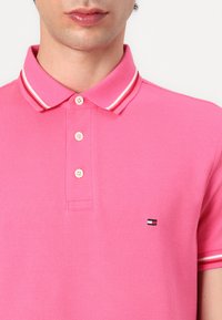 Rosa polo skjorta med texturerat tyg, tre knappknäppning och krage med vit-röda ränder. Har en liten logo på bröstet.