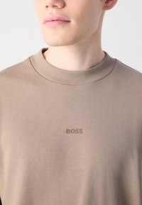 Sudadera de cuello redondo beige, fabricada con tejido texturizado, presenta un logo en relieve de "BOSS" en el pecho. Diseño clásico con cuello acanalado.