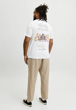 Mand med dreadlocks iført hvid t-shirt med blomsterprint, beige bukser og sorte sneakers, står med ryggen til mod en ensfarvet baggrund.