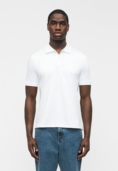 Hombre de pie de frente, vistiendo una camisa tipo polo blanca de manga corta y jeans de mezclilla azul sobre un fondo liso.