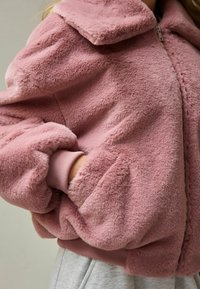Lyserød faux fur jakke med krave, ribbede manchetter, lynlås og blød tekstur, vist over et gråt tøj.
