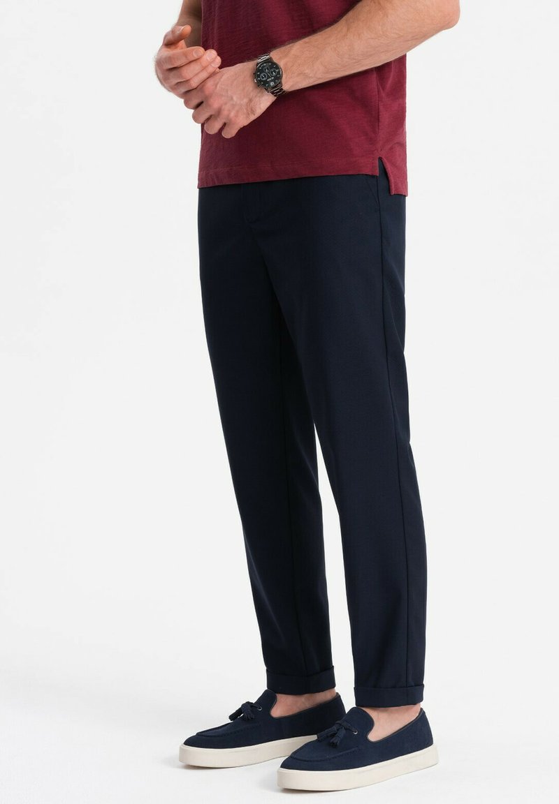 Pantaloni blu navy con una vestibilità sartoriale, caratterizzati da una texture liscia e orli arrotolati, abbinati a una maglietta bordeaux e scarpe slip-on blu navy.
