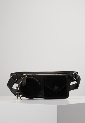 Sac banane en cuir noir avec une pochette ronde pour pièces, une poche carrée avec bouton, une fermeture éclair dorée et une sangle réglable sur surface blanche.