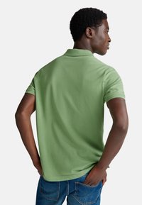 Zelené bavlněné polo tričko s klasickým límcem a krátkými rukávy, které má volný střih a hladkou texturu. Nosené s modrými džíny.