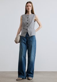 Γκρι μπλούζα χωρίς μανίκια με κουμπιά, συνδυασμένη με άνετο μπλε τζιν. Το μοντέλο κρατά μια τσάντα-πουγκί κρεμ χρώματος και έχει μακριά, ίσια μαλλιά.