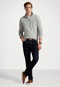 Sweatshirt cinzento claro com fecho e capuz, jeans de ganga escura, sapatos castanhos, cinto castanho e relógio de pulso. Ajuste casual; tecido parece macio e texturizado.