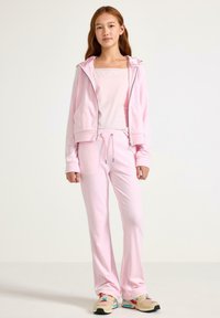 Jasnoróżowy dres z miękkiego materiału, obejmujący bluzę z kapturem i zamkiem oraz kieszeniami, spodnie ze ściągaczem i kolorowe buty sportowe.