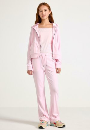 Tuta rosa chiaro in materiale morbido, con felpa con cappuccio e zip con tasche, pantaloni con coulisse e scarpe sportive colorate.