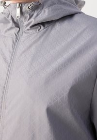 Veste à capuche gris clair avec motif géométrique diagonal, fermeture éclair frontale et cordons réglables près du cou.