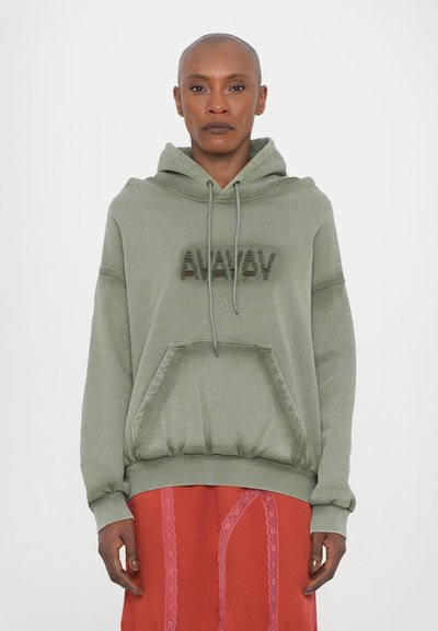 Sweatshirt à capuche vert clair avec une poche avant et un logo texturé. Manches retroussées et coupe décontractée, associé à une jupe rouge avec des détails en dentelle.
