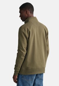Sudadera de algodón verde con cuello alto, puños acanalados y dobladillo, vista desde la parte de atrás. Combinada con jeans azules. Textura suave.