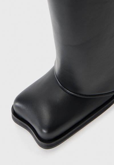 Botte de cheville en caoutchouc noir avec un bout carré, texture lisse et un léger talon. Détail de couture sur le côté et design minimaliste.