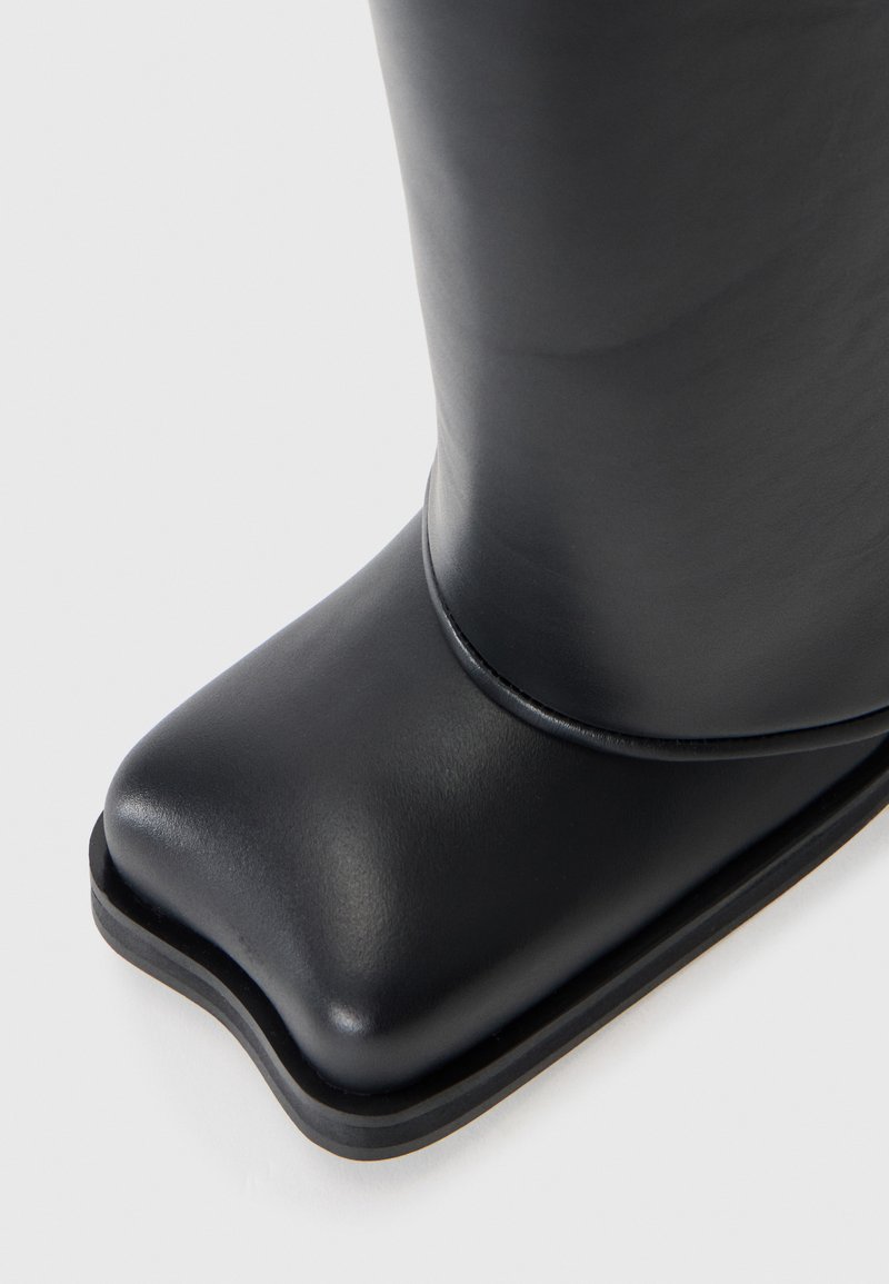Botte de cheville en caoutchouc noir avec un bout carré, texture lisse et un léger talon. Détail de couture sur le côté et design minimaliste.