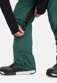 Hände, die die Beinte vents von grünen Outdoor-Hosen aufreißen und das schwarze Mesh-Innenfutter zeigen, kombiniert mit schwarz-weißen Wanderschuhen auf weißem Hintergrund.