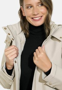 Bežinė vandeniui atspari striukė su auksinėmis detalėmis, užtrauktuku ir minkšta vidine pamušalu, derinama su juodu golfu.