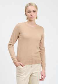 Maglione beige a coste con maniche lunghe e scollatura rotonda, caratterizzato da una consistenza morbida e una leggera lucentezza. Abbinato a pantaloni color crema.