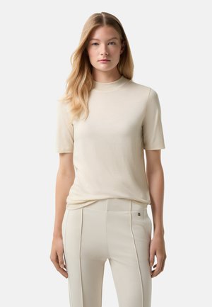 Beige kurzärmliges Oberteil mit hohem Kragen, aus glattem, leichtem Stoff gefertigt. Getragen mit hellen, körperbetonten Hosen.