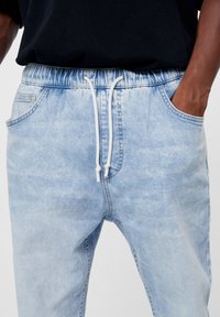 Světle modré džínové joggers s elastickým pasem, bílou šňůrkou a bočními kapsami. Obsahují jemné vyblednutí a mají uvolněný střih.