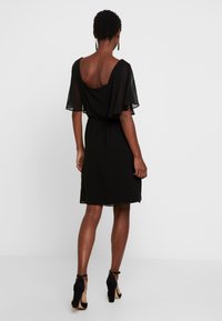 Black dress with short, sheer flutter sleeves, a scoop back, and an elastic waist. Smooth texture, knee-length design. Heels visible.