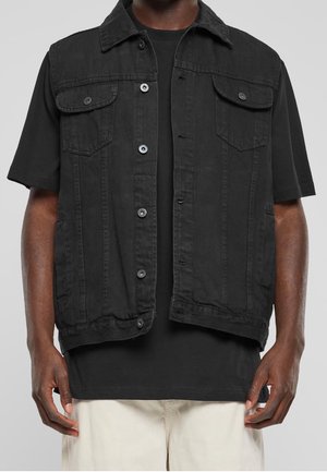 Person trägt eine schwarze ärmellose Jeansjacke mit Knöpfen über einem schwarzen T-Shirt und beigen Hosen, steht mit entspannten Armen.